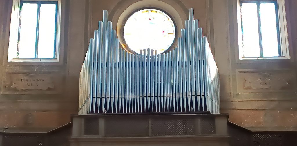 Huge church organ in front of a round window on a sunny day. Many pipes.