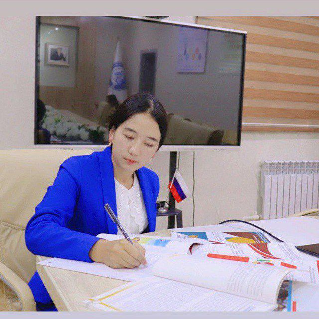 Young Central Asian woman with short black hair and a blue jacket and white blouse sitting at a desk writing. 