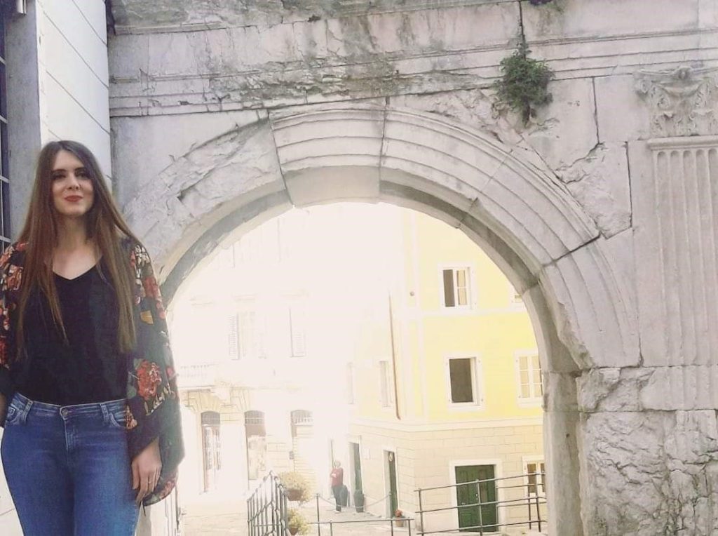 European woman with long brown hair, a black top, a sweater with roses, and blue jeans posing in front of a stone archway to a historical city. 