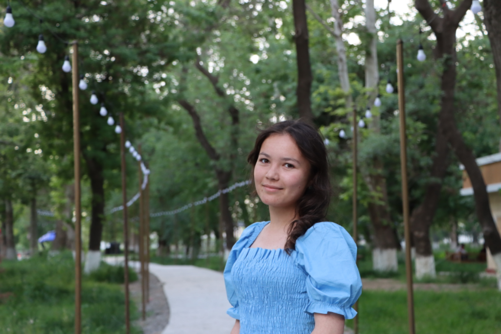 A Central Asian woman with black hair and a blue blouse standing in a park with trees and a trail behind her. 