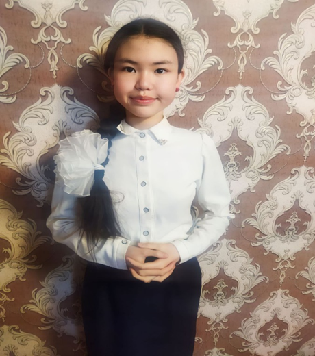 Young Central Asian girl with a white collared blouse and black pants and long black hair posing in front of a white and tan background.