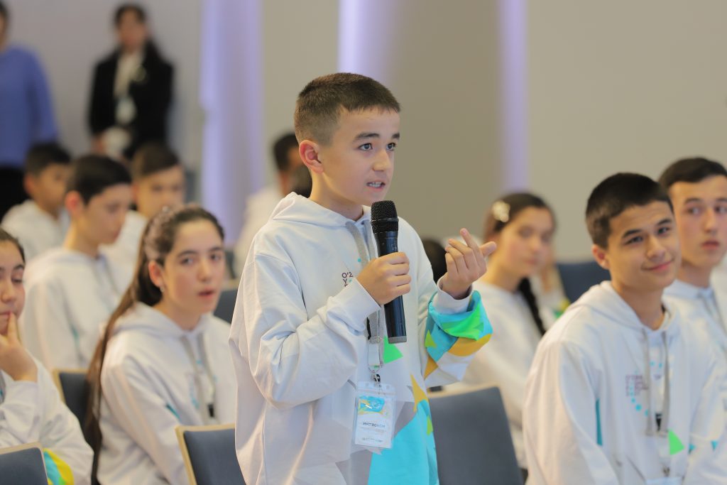 Young Central Asian boy stands up with a microphone in front of a group of other children, all dressed in white sweaters with colorful sleeves. 
