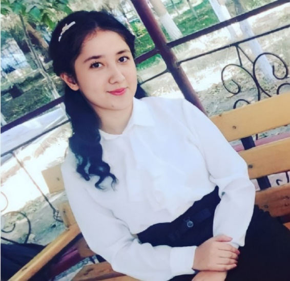Young Central Asian woman with curly black hair and a white collared shirt and black pants sitting on a park bench with a fence and trees in the background.