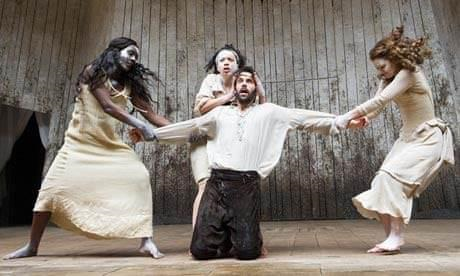 Theater production of Macbeth. Three young white women in white dresses and socks dance around and pull on the arms of a kneeling white man in a white shirt and black pants. 