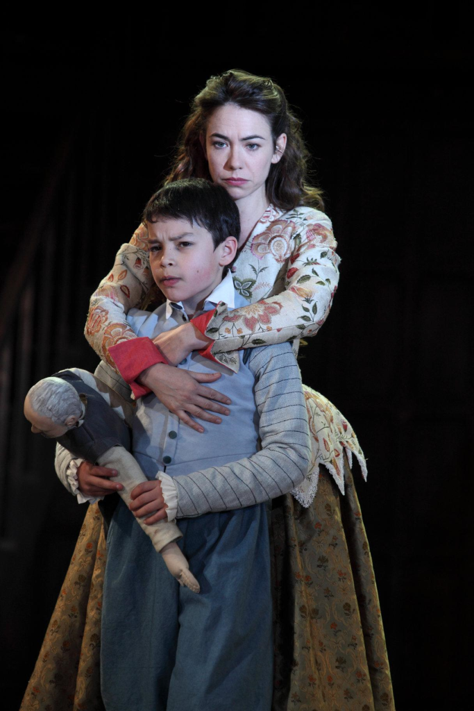 Macbeth's victims, Lady Macduff and her son. White woman with dark hair and floral shirt stands proudly embracing a small white boy with brown hair and a blue collared shirt holding a doll. 