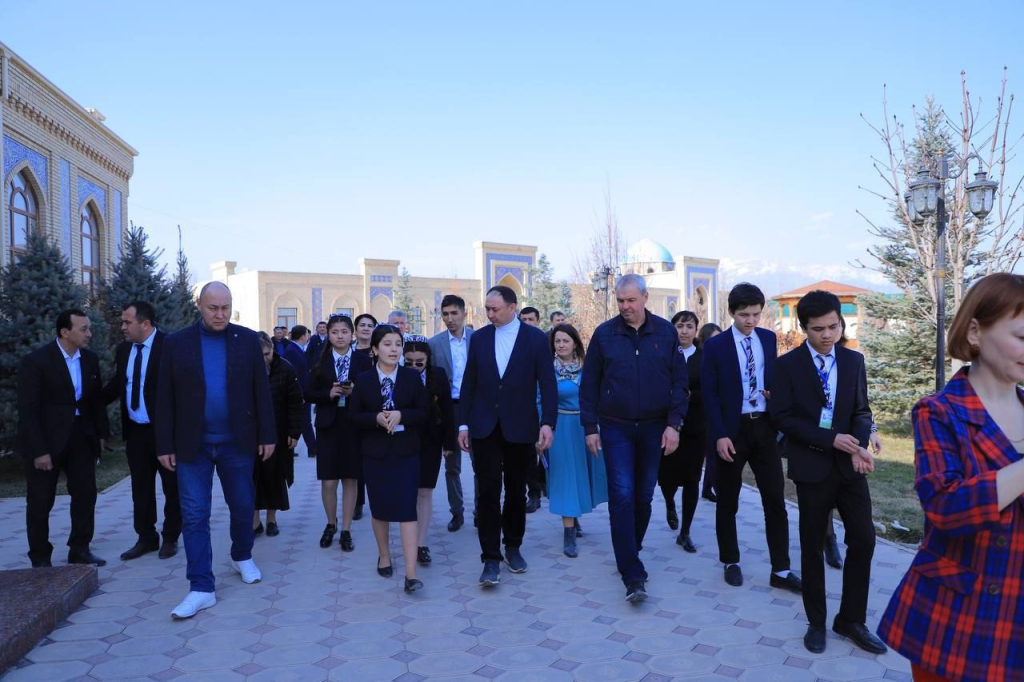 Uzbek education minister Boshkortostan and his colleagues escorted by Uzbek students.
Many men and women in suits and women in coats and blouses. Adults and high school kids walking on a concrete path outside of large school buildings. 
