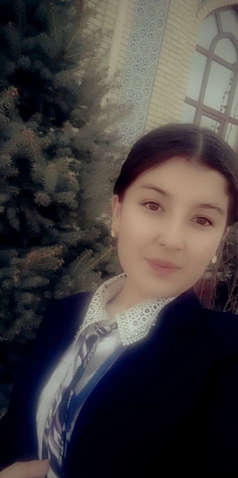 Young Central Asian woman with brown eyes and brown hair wearing a white blouse with a lacy collar and a black coat. She has earrings on and is standing in front of a large pine tree and a school building. 