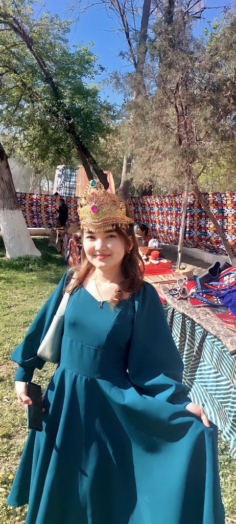 Young Central Asian woman with a crown on her head standing outside in a flowing blue-green dress with a crown on her head. She's at some sort of exhibition, out on a lawn where there are tables showing off jewelry and colored cloths. 