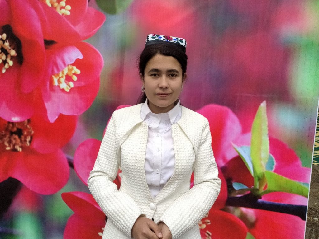 Young Central Asian woman with dark hair, a kerchief on her head, and a white knit jacket over a white blouse. She's in front of a bunch of pink and yellow flowers. 