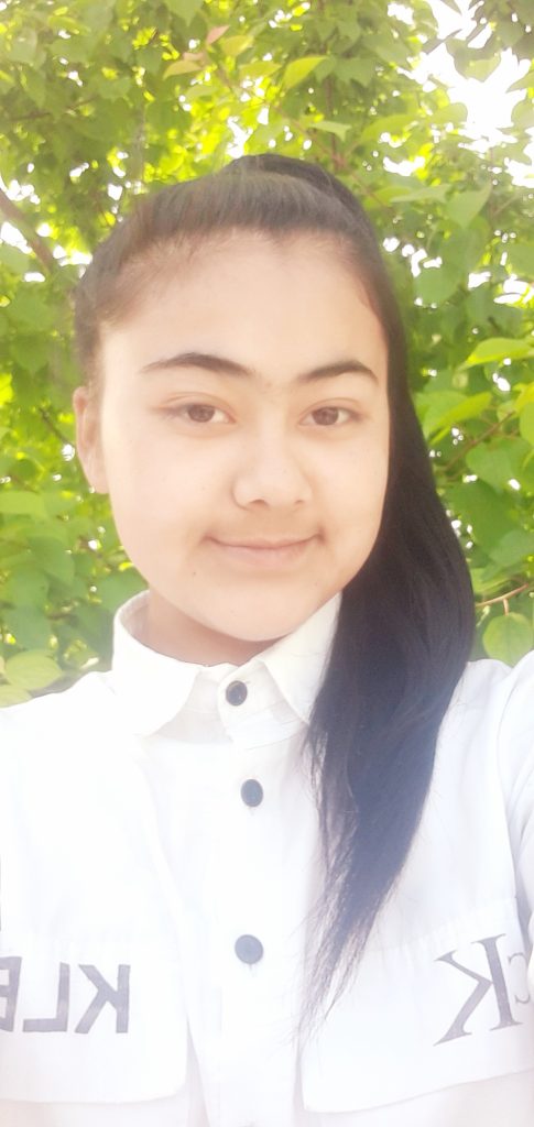Central Asian teen girl with long black straight hair up in a bun and ponytail, and a white collared shirt standing in front of a leafy tree on a sunny day. 