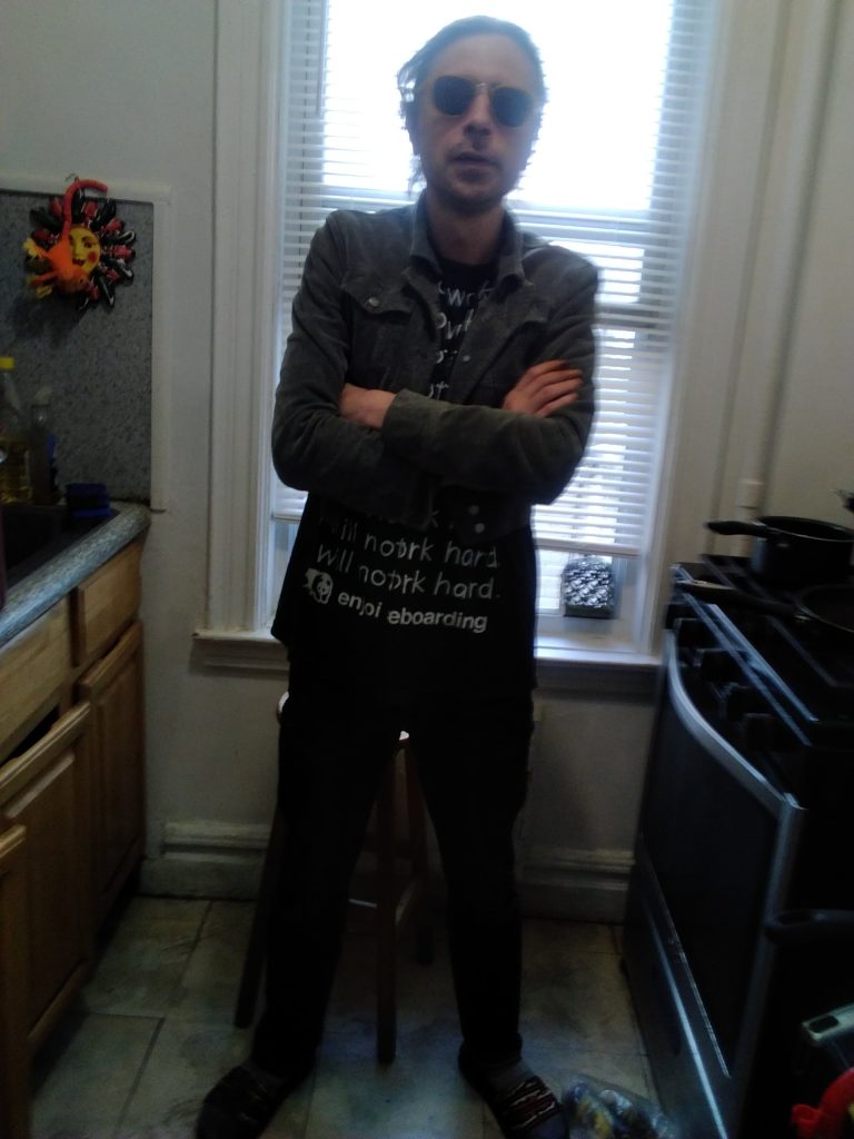Cool-looking white guy with sunglasses and short hair and a black jacket over a tee shirt with white lettering and black pants. He's standing in a kitchen in front of a window between the counter and the stove. 