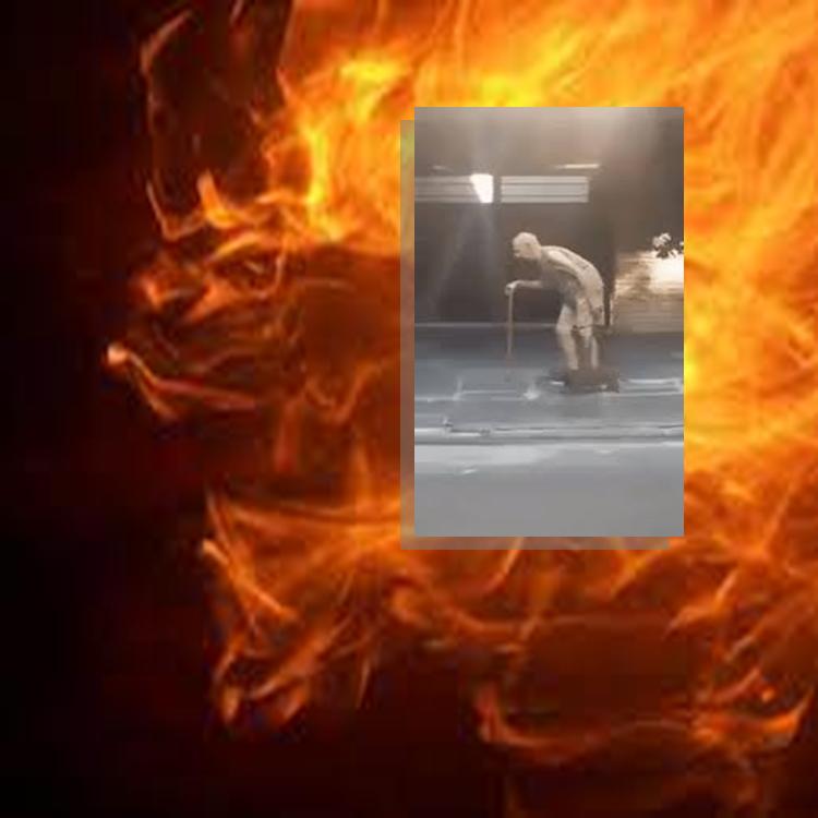 Fire in the background, image of an old man walking at night hunched over a cane in the foreground. 