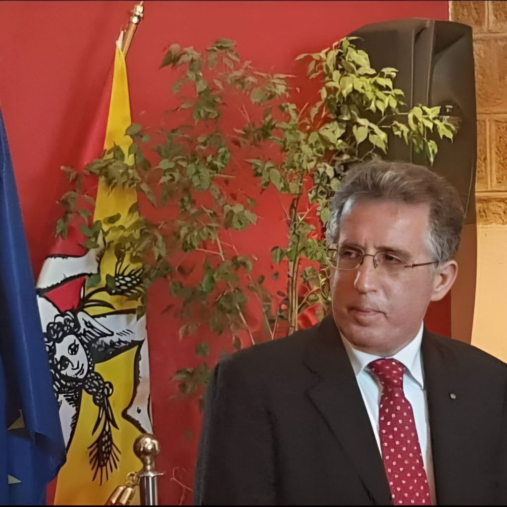 White man with short brown hair and glasses in a black suit and red tie standing in front of a red wall, a red and yellow and white flag, and a small houseplant tree. 