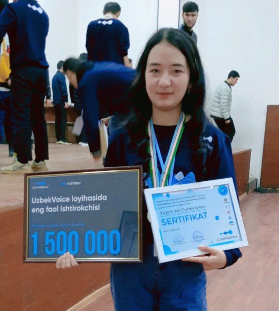 Young Central Asian teen with dark hair and brown eyes stands in a roomful of fellow students holding a certificate. 