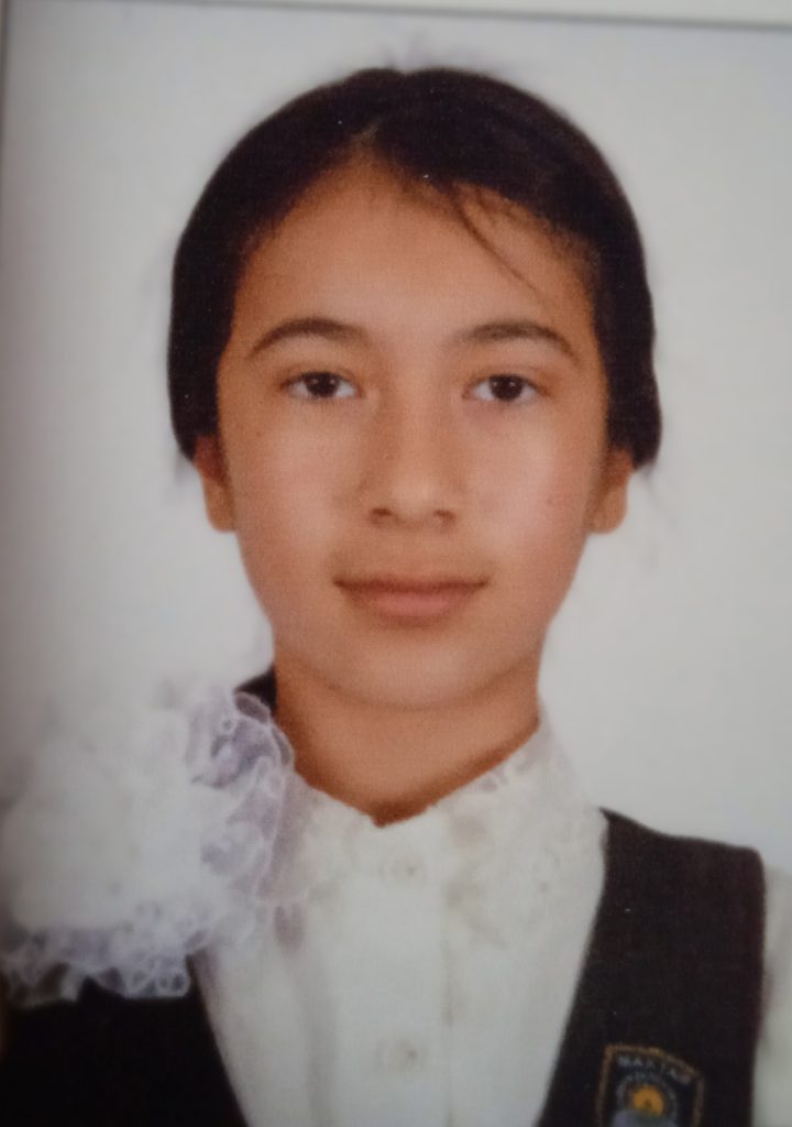 Young Central Asian woman with brown hair pulled behind her back, brown eyes, a white buttoned blouse with a white flower, and a black vest with an emblem on the right. 