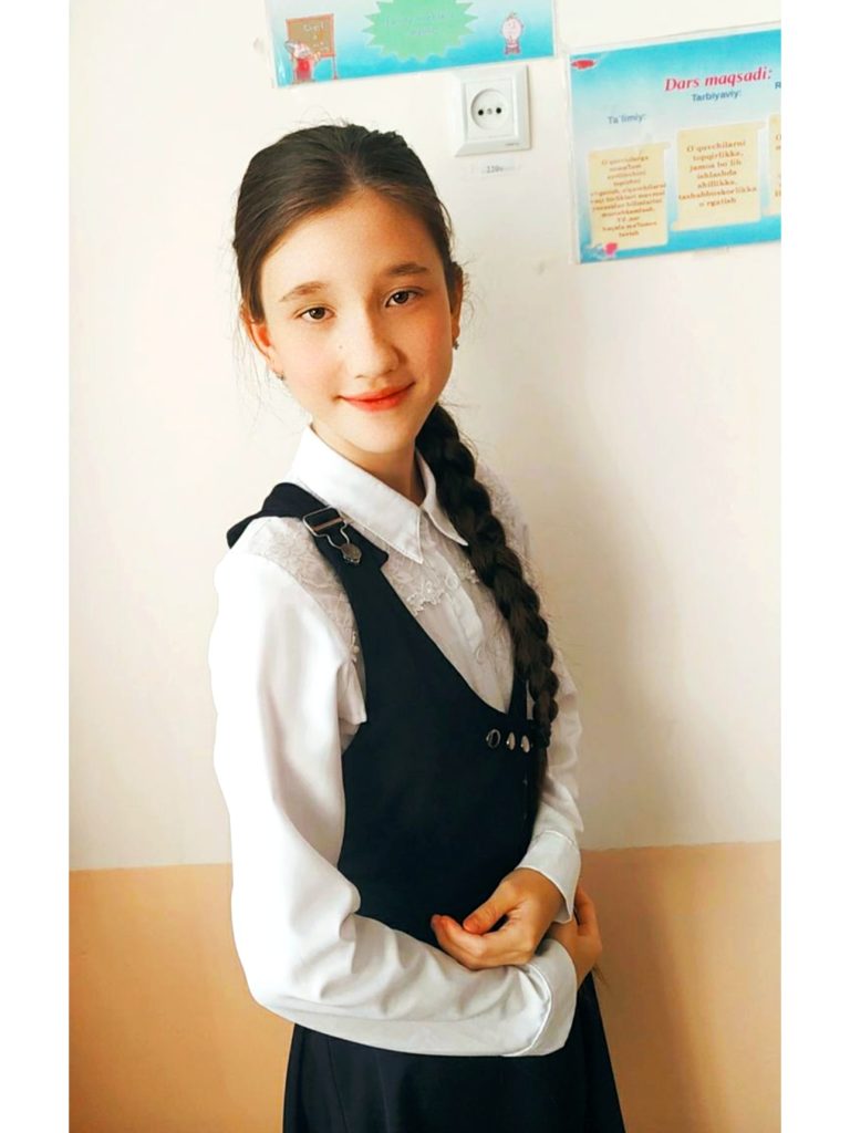 Young Central Asian girl with a long black braid, brown eyes, and a white collared ruffly top and black overalls. 