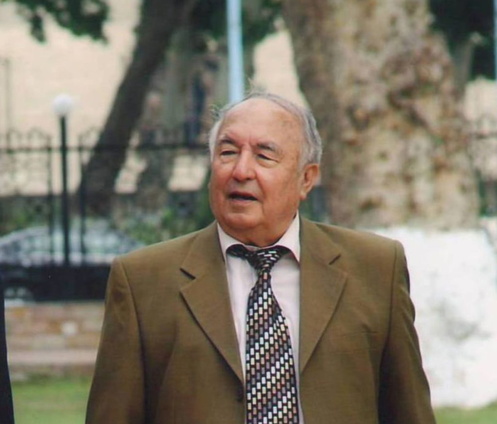 Older Central Asian man with grey hair, a tie, and a white shirt and brown coat standing outdoors in a park with a black iron fence and a tree. 