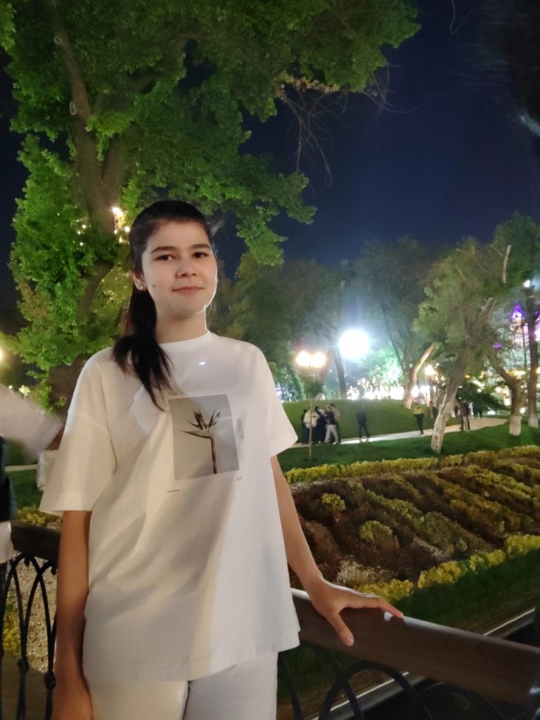 Young Central Asian girl in a tee shirt with a flower on it in black and white. She's in a park at night in front of a fence, flowers, grass and a hiking path.