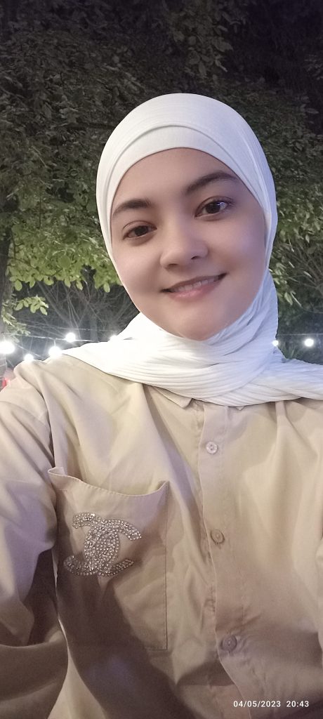 Young Central Asian woman with a white headscarf and a peach buttoned shirt. She's standing outside in the evening in front of leafy trees. 