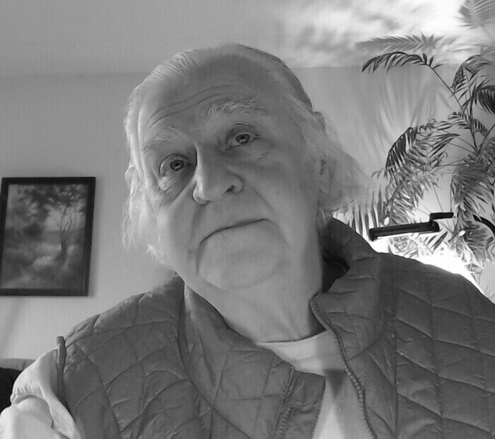 Older white man with a coat and a tee shirt in his living room with a houseplant in the background and a picture on the wall.