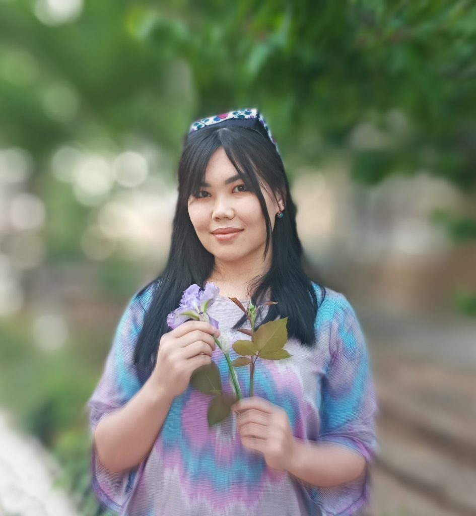 Young Central Asian woman with straight long black hair and a headband holding a purple flower. She's standing in front of a blurry background of trees.