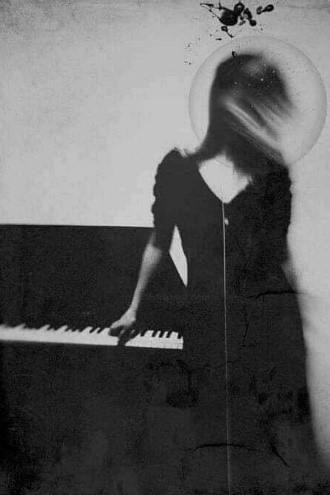 Woman with a halo around her hair facing a wall and touching a piano. She's in a long black dress. 