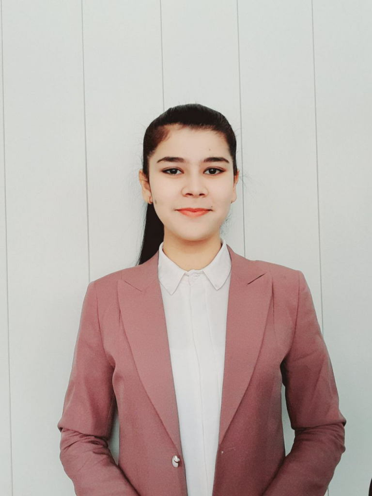 Young Central Asian woman with long straight brown hair behind her head, brown eyes, a beige collared shirt and pink jacket standing in front of a white paneled wall. 