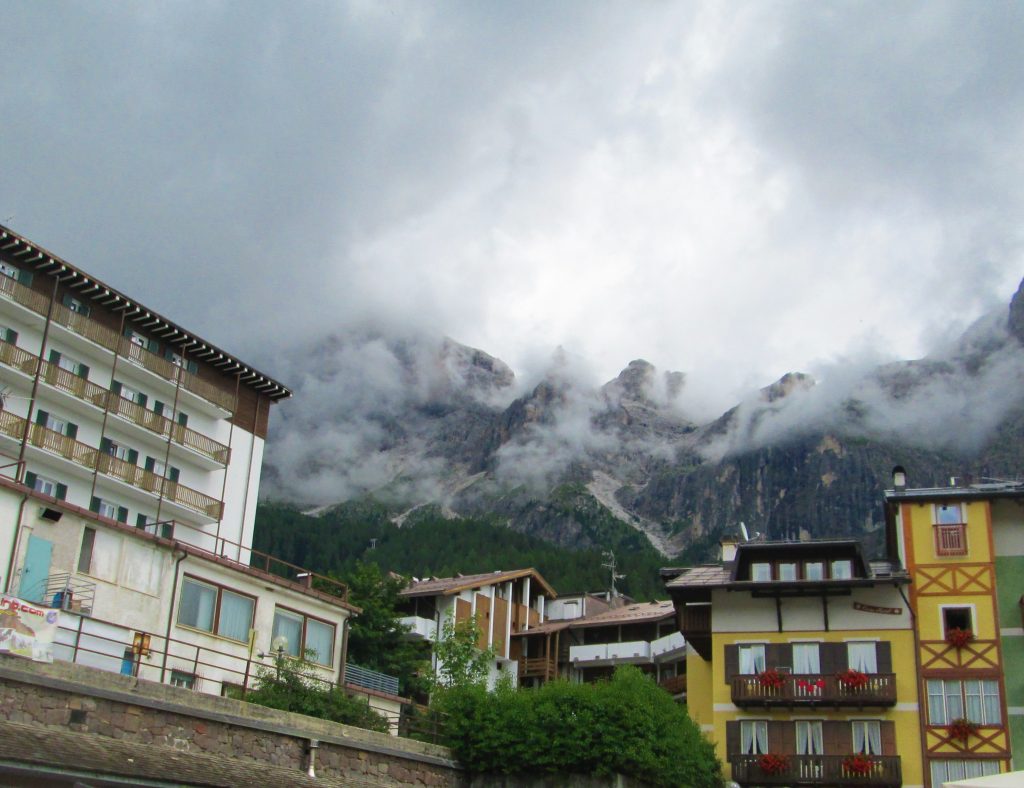Multi-story buildings in yellow, white, and brown in front of large mountains with clouds and trees. 