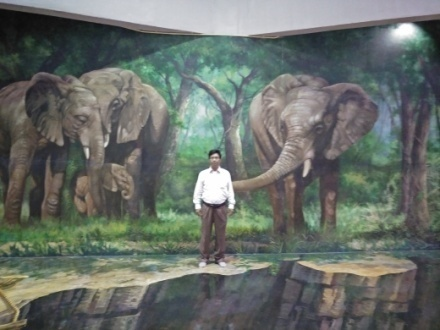 South Asian man in white shirt and brown pants standing in front of a giant painting of elephants. 