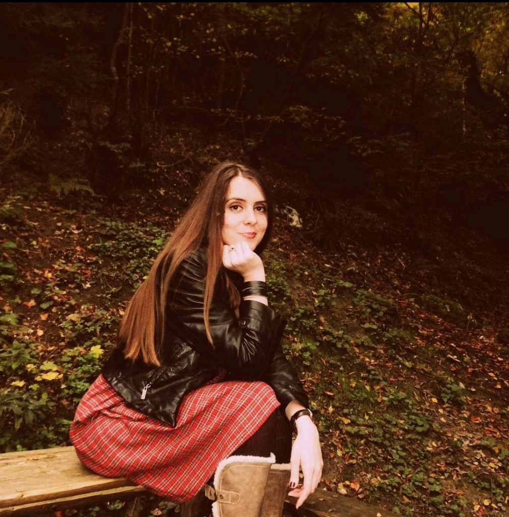 Young white woman sitting in a field. Dark area, twilight or just a lot of thick trees behind her. She's got long brown hair and a black leather jacket and a red plaid skirt and brown furry boots. 