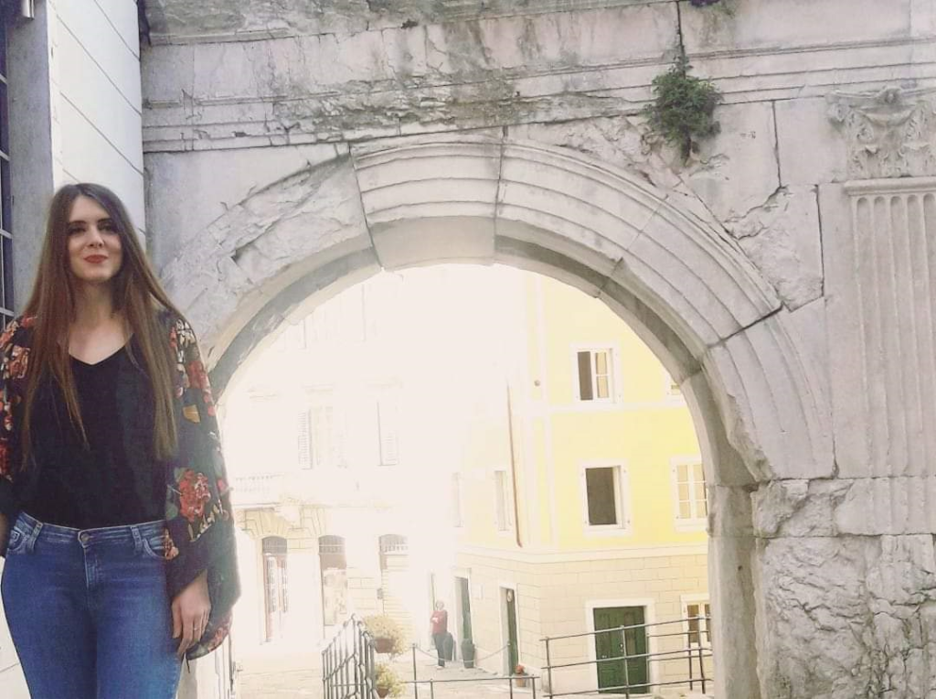 Young white woman stands to the left of a picture with brick ruins and an arch in the background. She has waist length dark hair, a black top and blue jeans, and a floral jacket.