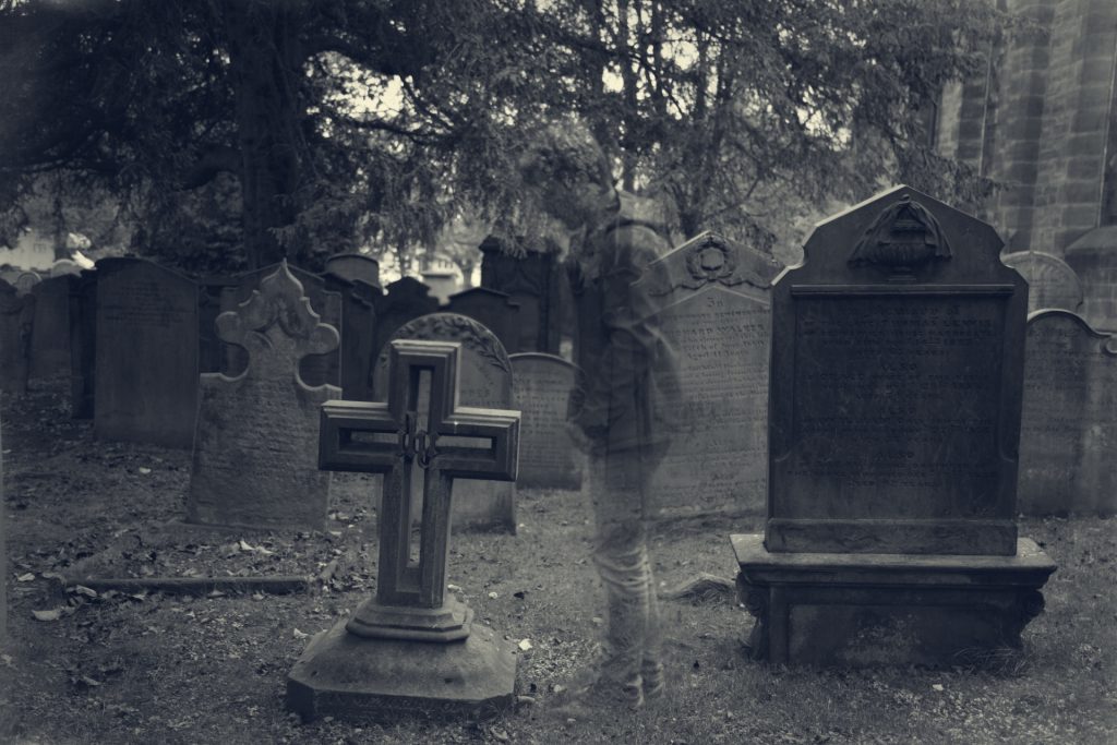 Ghostly human figure, a person with a coat and jeans and tennis shoes, stands in front of gravestones and crosses in a cemetery. 