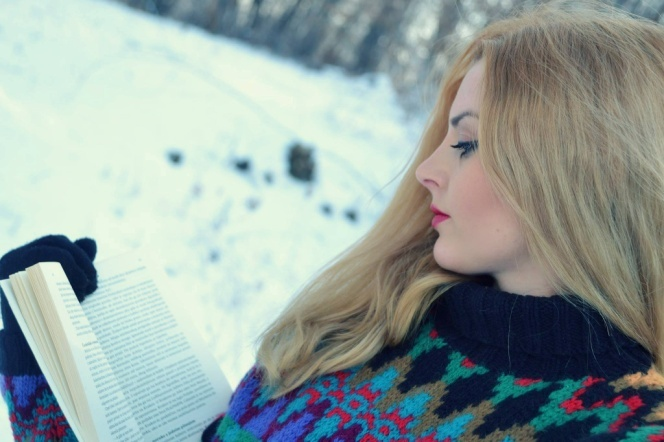 White woman with long reddish hair reads a book outside in the snow. She is wearing a colorful sweater. 