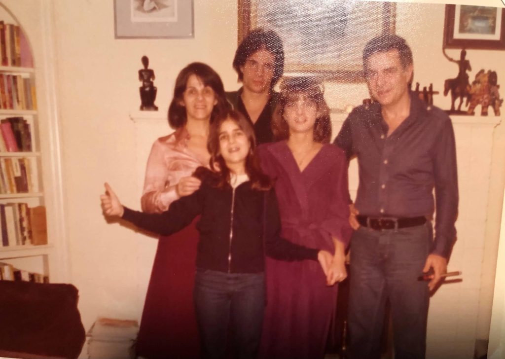 A man and a woman and a teen boy and two girls stand in a living room with paintings and a bookshelf in the background. Guys wear collared shirts and jeans and belts, women and girls wear red dresses, except the youngest girl who has jeans and a black jacket on. This is a photo of an old-time physical photograph. 