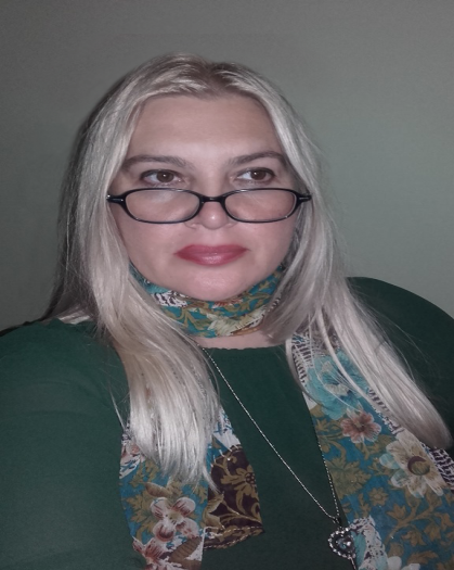 Younger middle aged white woman with long blonde hair, glasses, and a green top and floral scarf and necklace.