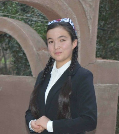Young Central Asian woman with long braided black hair, a headband, and a white blouse and black jacket. She's in front of trees and stone arches. 