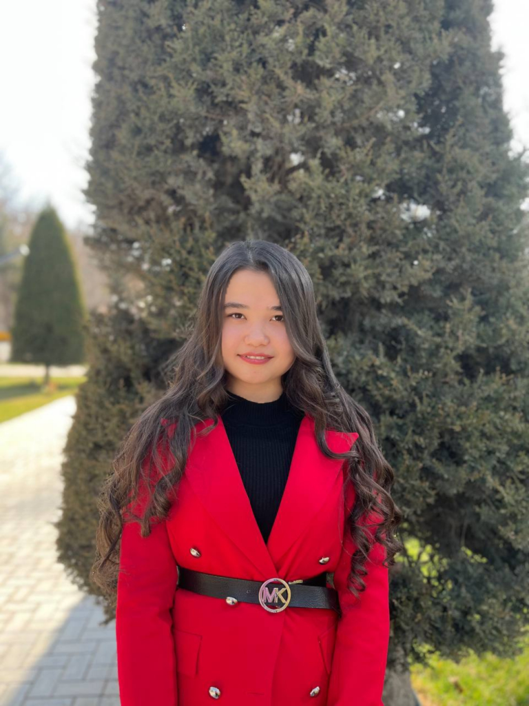 Young Central Asian woman with long curly black hair and a black top and red jacket and brown belt standing outside on a brick sidewalk in front of a tree and lawn. 