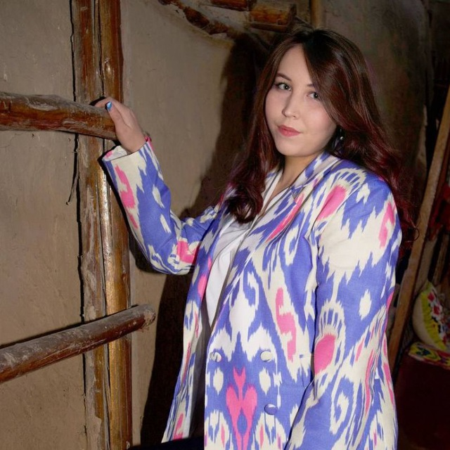 Young Central Asian woman with long brown hair and a colorful pink and purple jacket over a white blouse standing in a room next to a ladder.