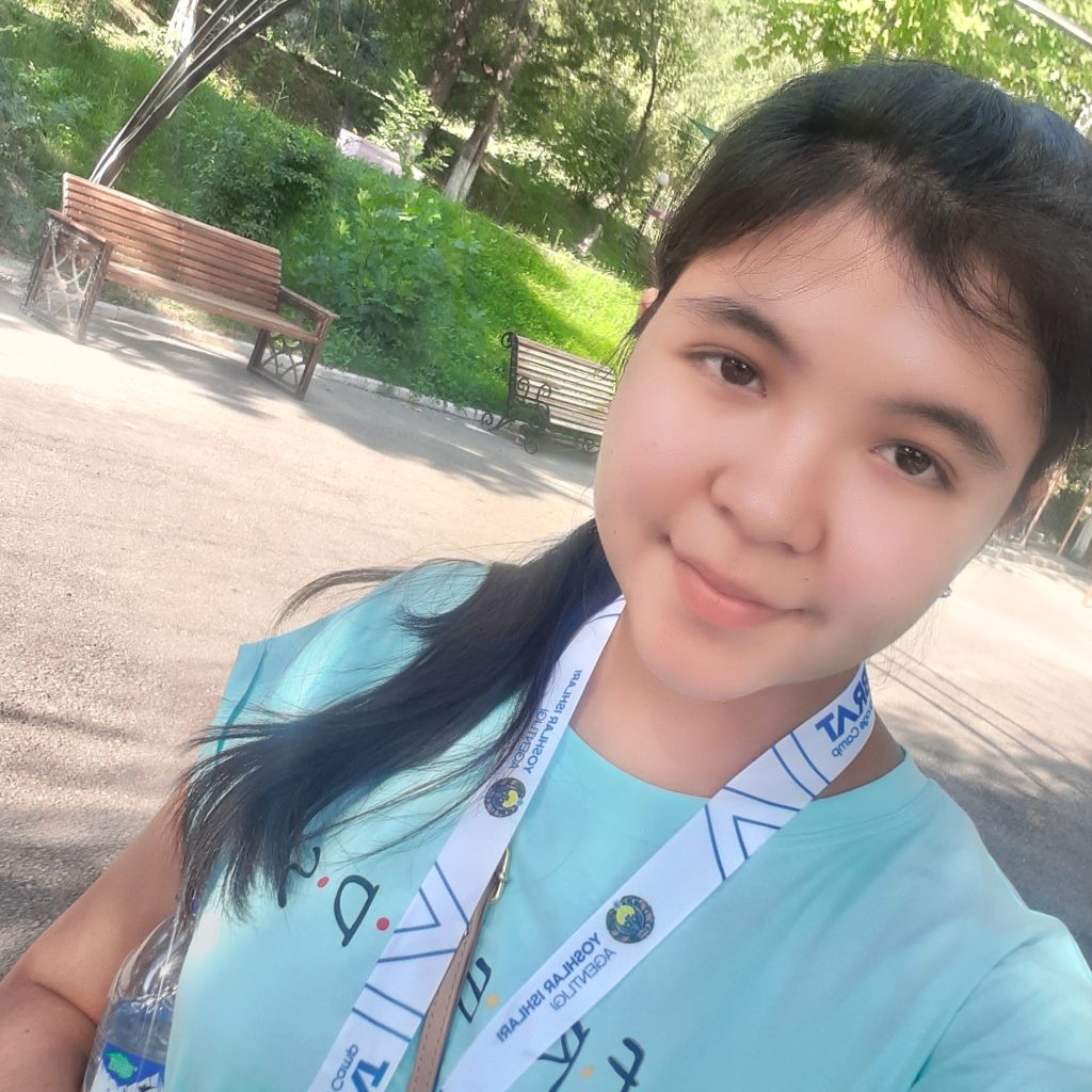 Young Central Asian teen with long black hair and a lanyard around her neck and a pale blue tee shirt. She's outside by some park benches and a lawn and on a gravel path. 