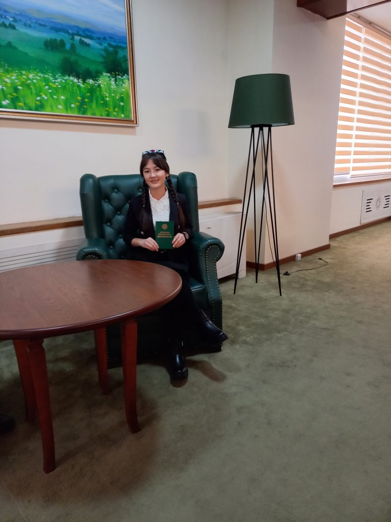 Young Central Asian woman with braids and a white top sitting down and holding a green book. She's in a large velvet poofy chair in a living room with a lamp and carpet and table and picture on the wall of a pastoral scene of rolling hills, clouds and trees. 