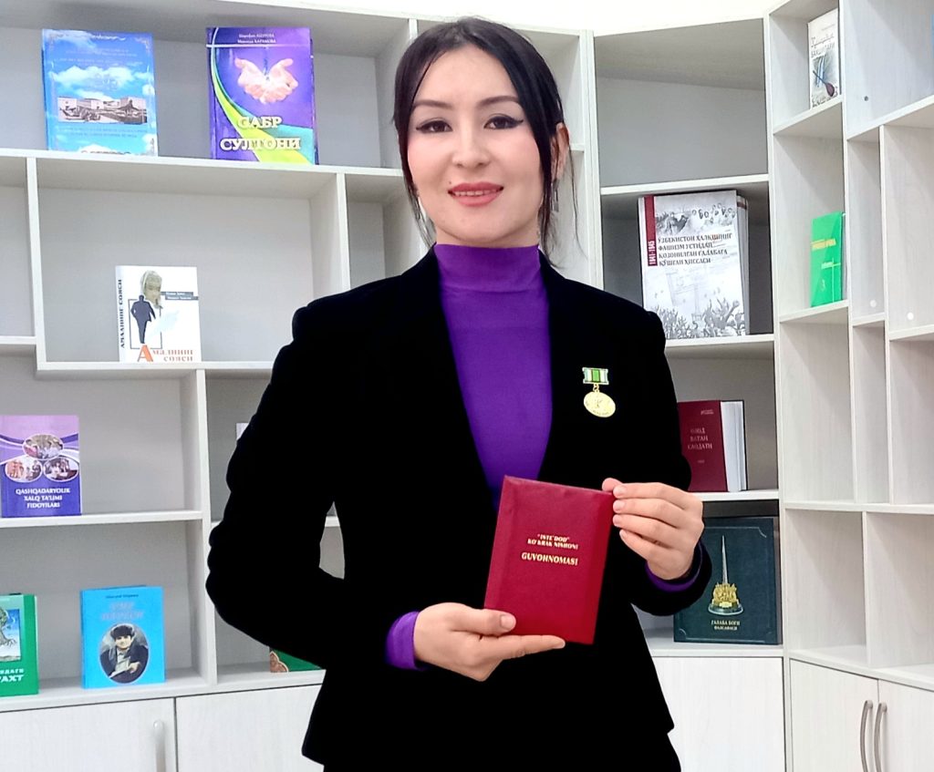 Young Central Asian woman with short straight dark hair, a purple turtleneck and a black jacket and a medal on her breast. She's holding a red certificate and standing in front of a bookshelf with several books on display. 