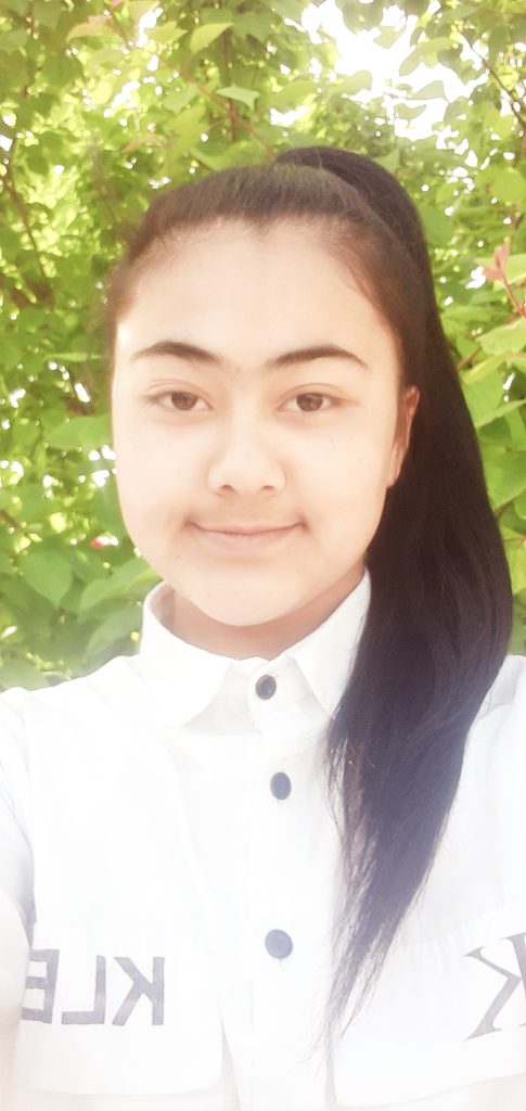 Central Asian teen girl with her hair in a ponytail and a white collared button down shirt. She's standing in front of a leafy tree. 