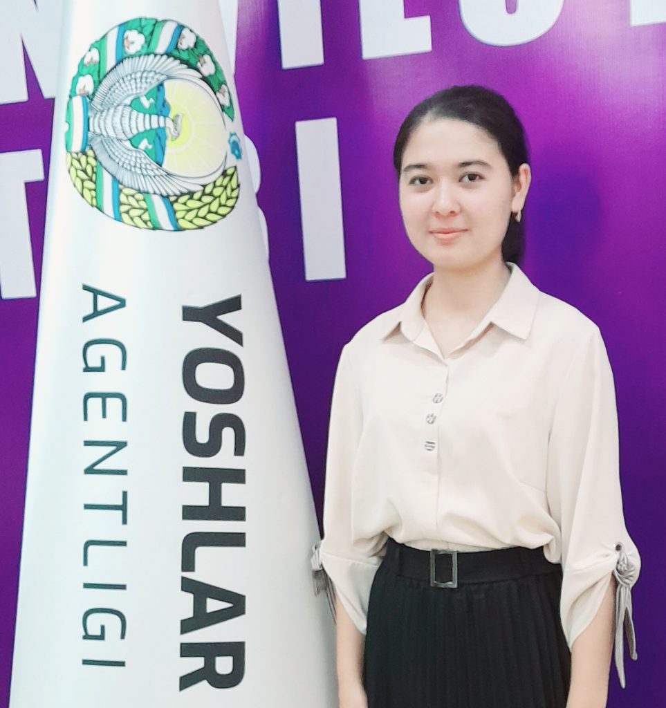 Young Central Asian teen girl with long dark hair behind her head and a off white collared shirt with buttons. She's in a black skirt with a belt and standing in front of a banner at an event on stage. 