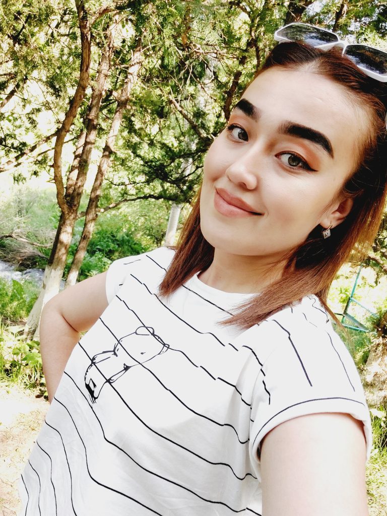 Young Central Asian woman with dark hair, earrings, brown eyes, glasses on top her head, and a striped white and grey shirt. She's standing outside by trees and grass and a brook. 