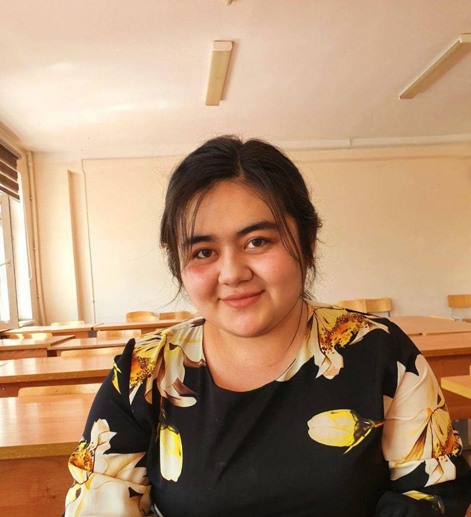 Young full figured Central Asian teen girl with dark hair and brown eyes in a black blouse with yellow flowers. She's in a classroom with wooden desks.