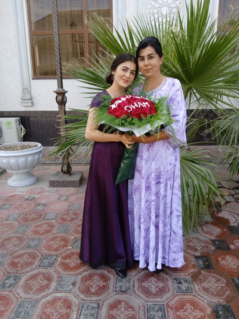 Young Central Asian woman with a dark purple dress standing next to an older Central Asian woman with a light purple dress standing in front of a house with a window and a palm tree. Both women are holding a bouquet of roses. 