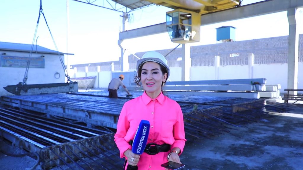 Young Central Asian woman in a pink collared shirt and a hat standing in a shipyard near wooden pallets and a yellow crane. 