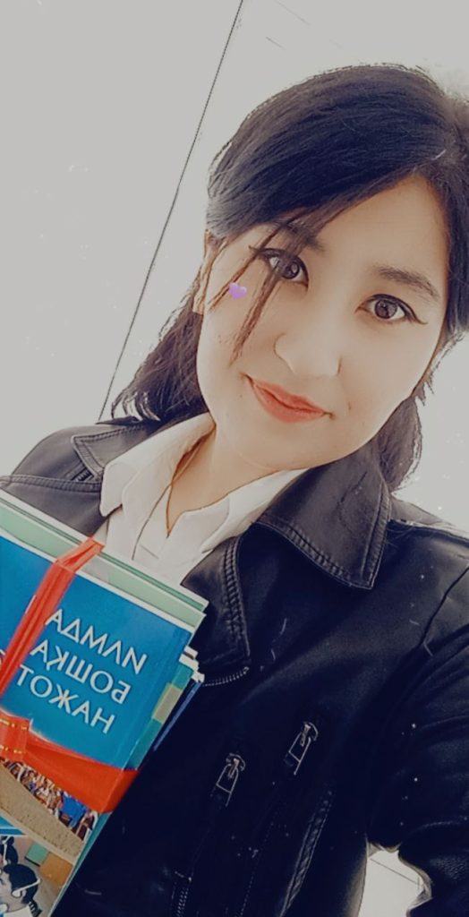Young Central Asian woman with long black hair and brown eyes and lipstick. She's got a white collared shirt and black jacket and is holding a textbook.