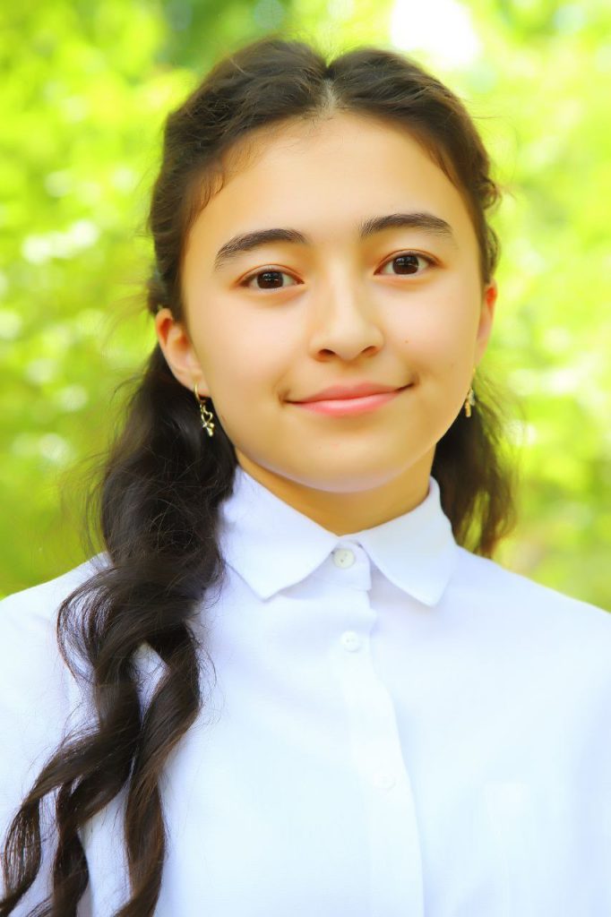 Young Central Asian teen girl with a slight smile and her hair up behind her head and earrings. She's in a white collared shirt and standing in front of leafy trees. 