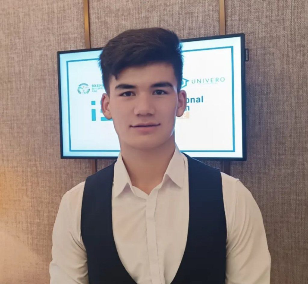 Young teen Central Asian guy with short dark hair, a white collared shirt, a black vest and a serious expression. He's standing in front of a screen with the logo of his school. 